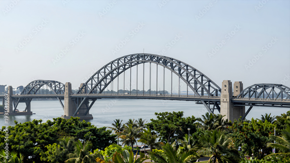 Fototapeta premium Large Steel Arch Bridge Spanning Wide River with Lush Green Palm Trees in Foreground under Clear Sky for Travel Architecture and Urban Development Concepts