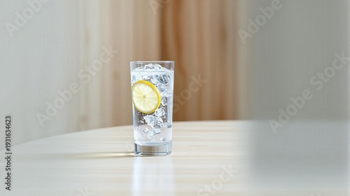 A tall glass of clear cool water filled with ice and a bright yellow lemon slice sits on a wooden table with a soft brown curtain in the background