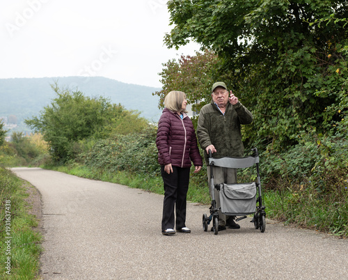 Elderly married couple taking a walk in the park with help of walker to support and help move