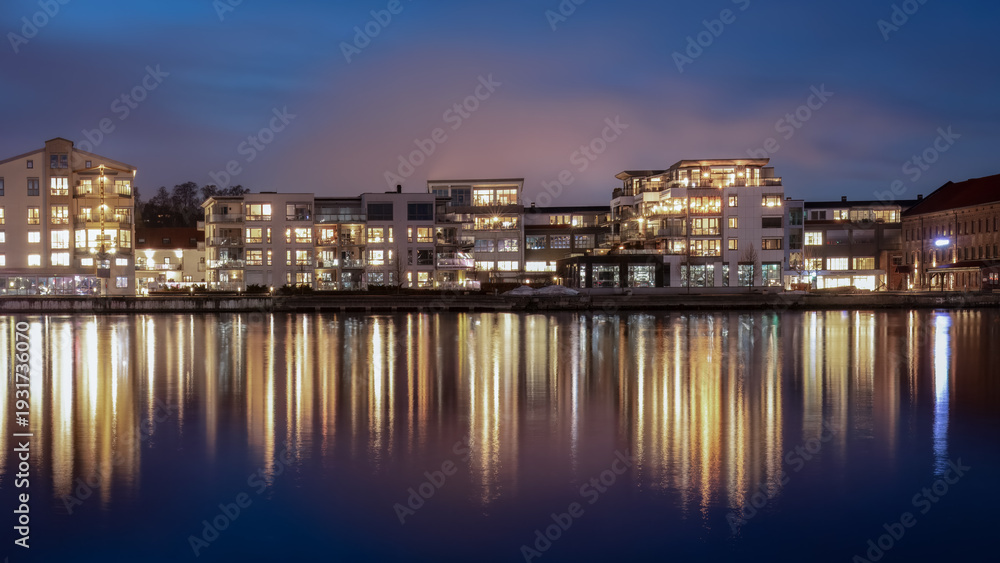 Obraz premium Authentic Scandinavian river culture illuminated residential waterfront in Porsgrunn Norway with glowing apartments mirrored in still river under northern evening sky highlighting local urban identity