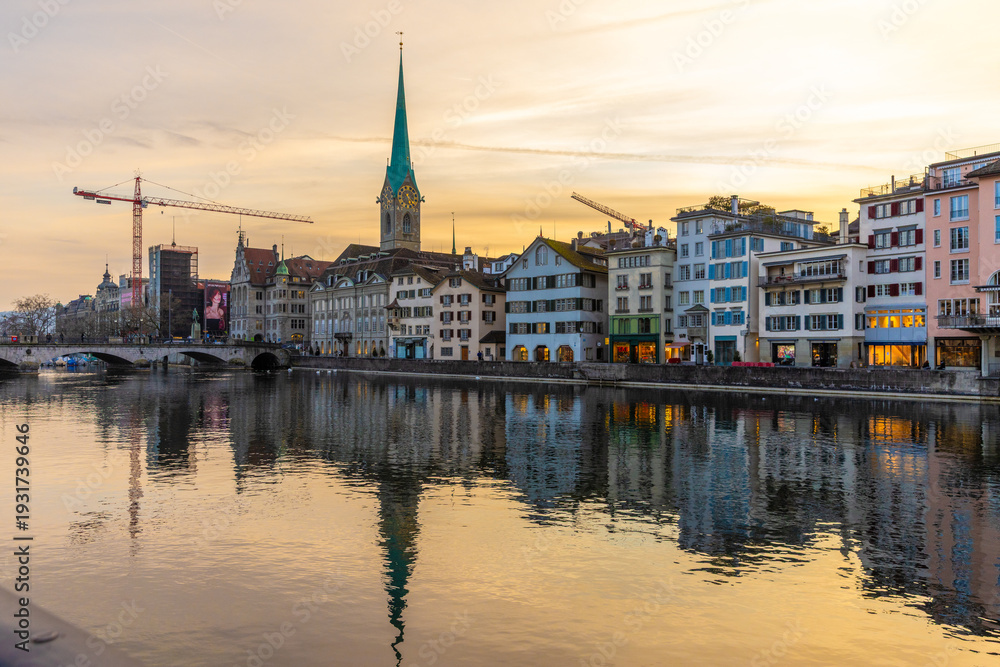 Fototapeta premium Panoramic evening view of the Limmat river in Zurich, Switzerland, showing colorful historic buildings and the Fraumünster church spire reflected in the golden water at sunset.