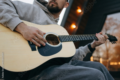 Foto Man plays acoustic guitar outdoors at dusk with warm lights and relaxed vibe