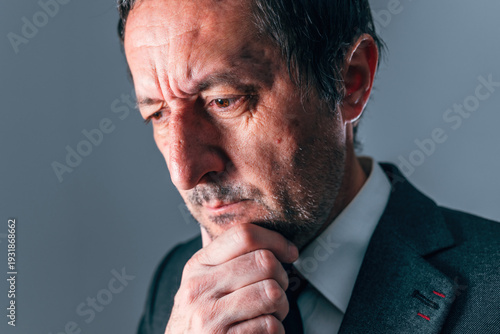 Portrait of a businessman in dark suit and tie, chin resting thoughtfully on his hand, filled with concern for deep reflection, decision making and inner conflict concepts.