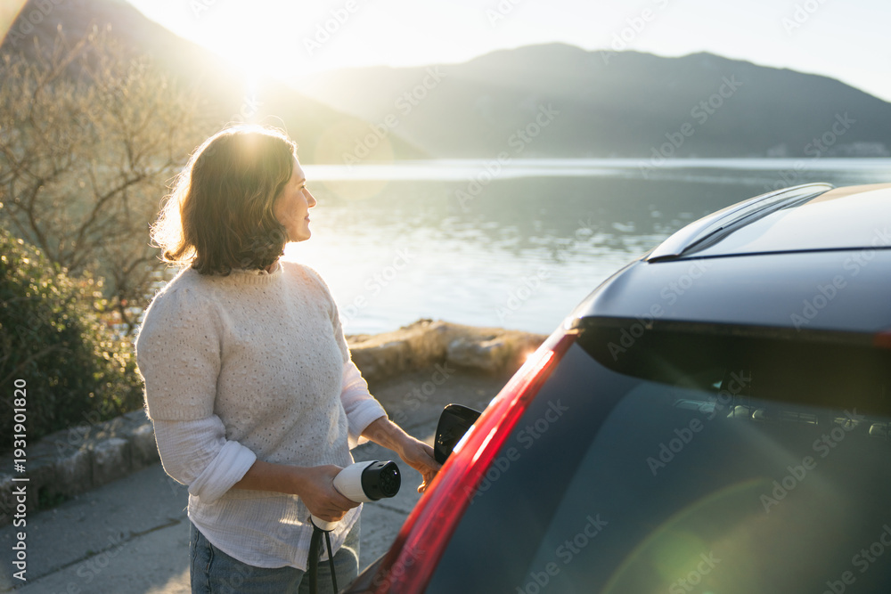 Fototapeta premium Woman traveler with charging plug next to her electric car on the sea coast at sunset