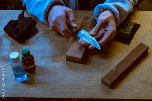 Close-up of hands testing the sharpness of a vintage knife blade over a workbench with sharpening stones and oil.