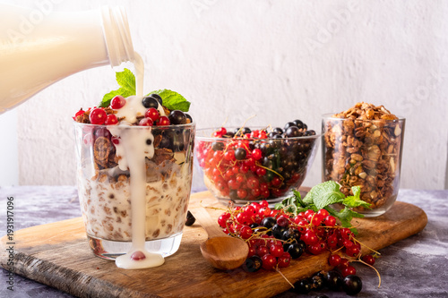 Pouring milk into granola glass with spilled drops, fresh currants, and nuts on a table.