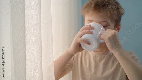 Boy at the window drinking milk, tea or cocoa from a white ceramic mug. Focus on child nutrition, dairy products, breakfast routine, and healthy beverage intake at home for kids.