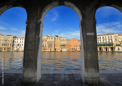 Scenic view of the Grand Canal in Venice framed by historic stone arches. Colorful Venetian palaces reflected in the water under blue sky. Iconic Italian architecture, travel destination and romantic 