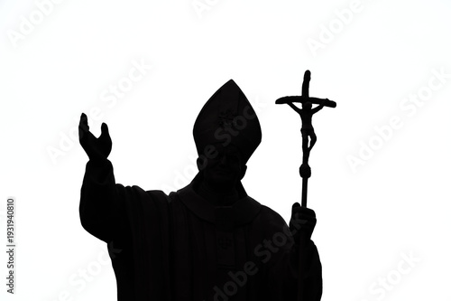 Photography A Catholic pastor in a cassock and cap with a cross in his hand, silhouetted against a light background