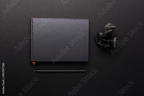 Black notebook with red bookmark, black pencil and crumpled paper on dark desk.