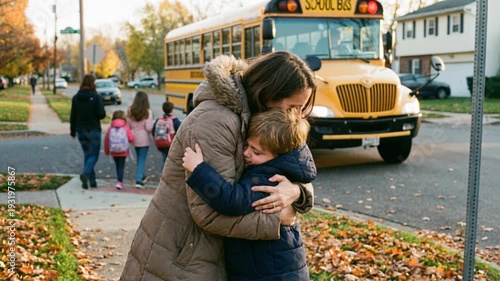 Woman and young boy hugging on sidewalk as children walk to school bus, autumn season and family relationships concept.