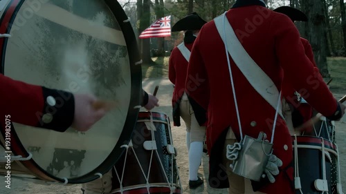 Colonial soldiers in red coats marching with drums and a flag in the background