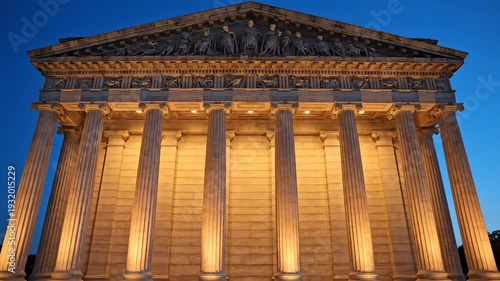 Neoclassical building facade with ionic columns illuminated by warm light, wide architectural shot at dusk with symmetrical composition, tall fluted columns and ornate entablature glowing under soft