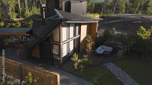 Aerial drone view of a modern luxury cabin with contemporary A-frame architecture, nestled in a lush forest with mountain views and a landscaped garden.