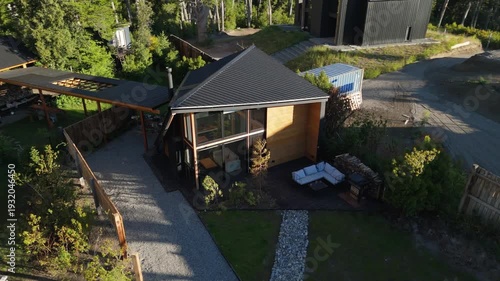 Aerial drone view of a modern luxury cabin with contemporary A-frame architecture, nestled in a lush forest with mountain views and a landscaped garden.