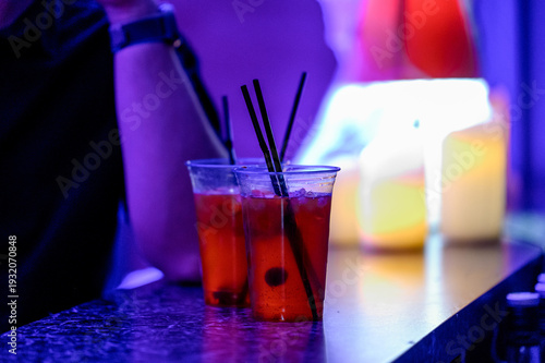 Two cocktails with straws on bar counter, illuminated by colorful lights