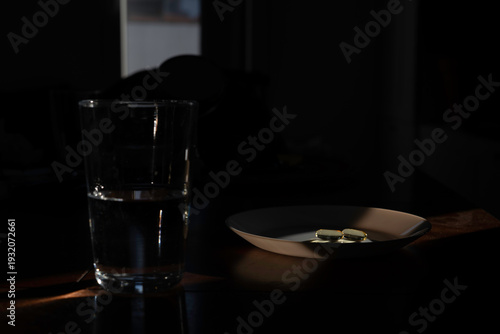 Pills and a glass of water are waiting on a table in a dark room