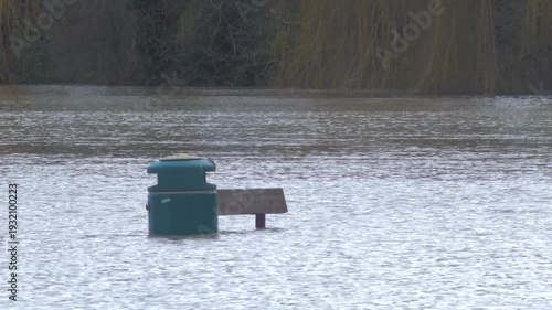 park bench and rubbish bin aka trash can in flooded city park, poking through the water, trees in background