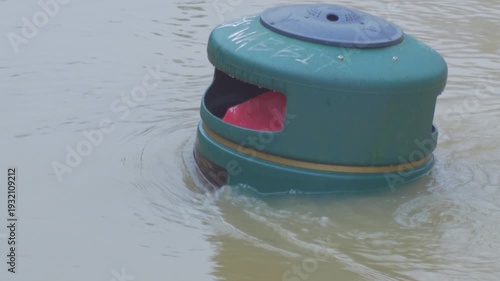 flood water nearly covering rubbish bin aka trash can, top of bin visible, surrounded by fast flowing muddy water, flooded city park