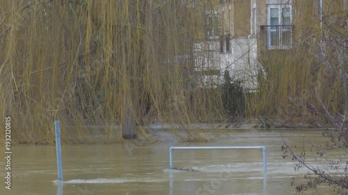 Flooded river, fast flowing muddy brown water, willow trees and residential apartments in background, submerged railing and signpost in foreground