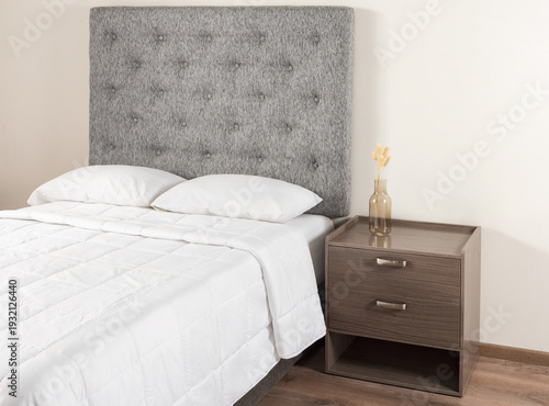 A bedroom featuring a bed with white bedspread and a grey fabric headboard, a nightstand situated beside it