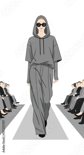 A female model in an oversized grey hoodie and wide-leg pants, wearing sunglasses, walks down a grey runway flanked by seated audience members.