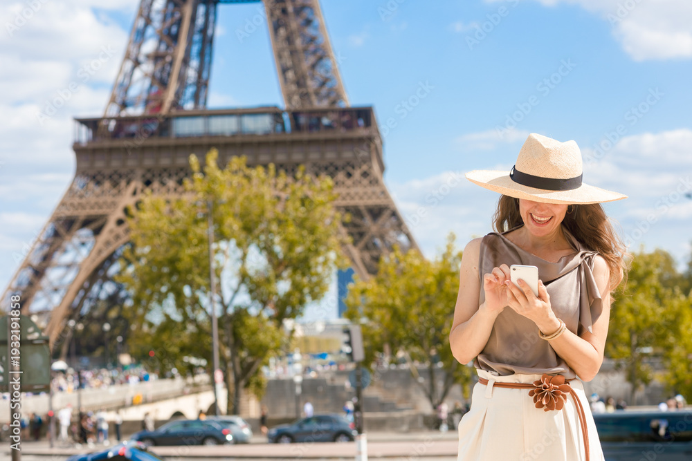 Ταπετσαρία A happy woman in a straw hat checks her smartphone, with the magnificent Eiffel Tower subtly blurred in the background, capturing a moment of connection during her Parisian vacation