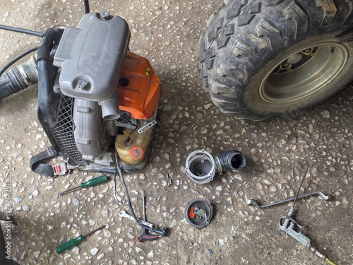 Disassembled orange leaf blower on workshop floor. messy repair project with various part and tool scattered around dirty tire. Work in progress in garage