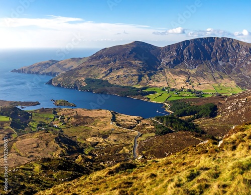A wide landscape showing a large lake bordered by mountains and the sea. The view is from a high vantage point. Clear skies