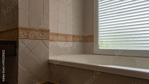 A white rectangular bathtub in the corner of the bathroom. Beige tiled walls. Decorative finishing strip with an ornament. Blinds on the window.