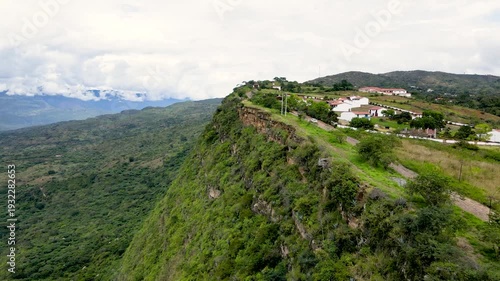 This aerial shot captures the lush green cliffs of Barichara, Santander, with white houses nestled along the ridge under a cloudy sky.