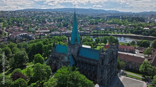 Aerial view of Nidaros Cathedral in Trondheim, Norway, Norwegian treetops surrounding the nave and transept . Drone view eases past the spire, opening the frame to the river bend and city blocks.