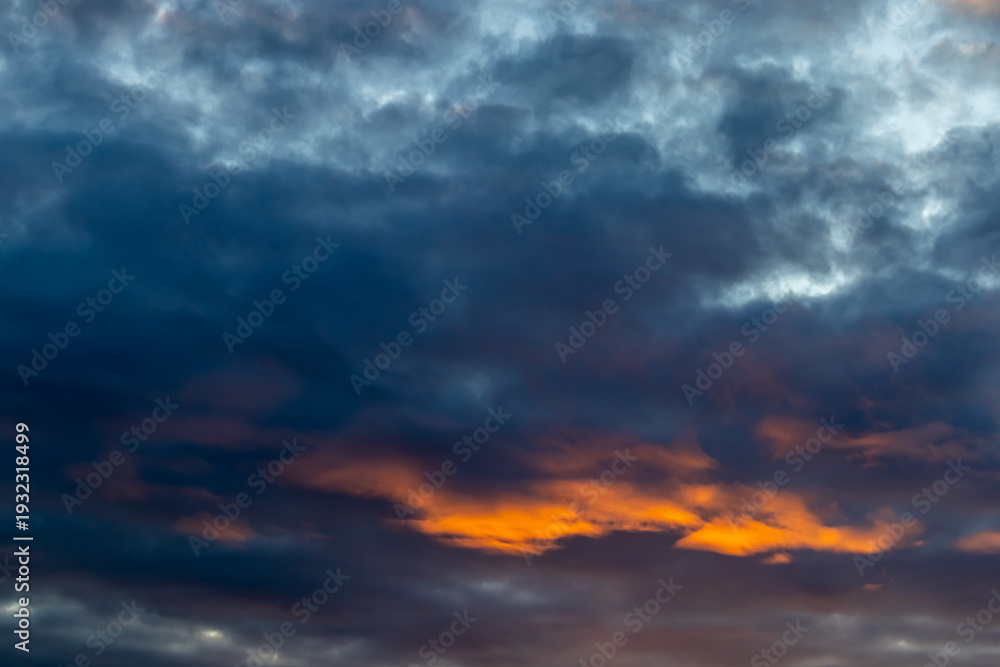 Obraz premium Dramatic view of a darkening sky where bright orange light breaks through thick layers of blue and grey clouds