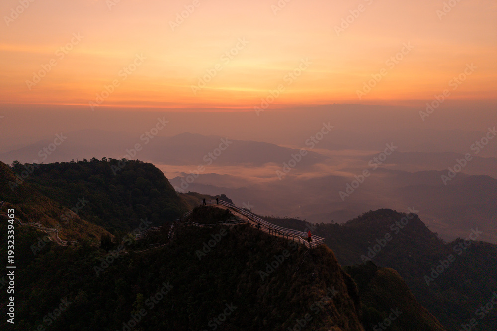 Obraz premium Aerial view of a mountain ridge path with a fence overlooking a breathtaking valley and sea of clouds at sunrise.