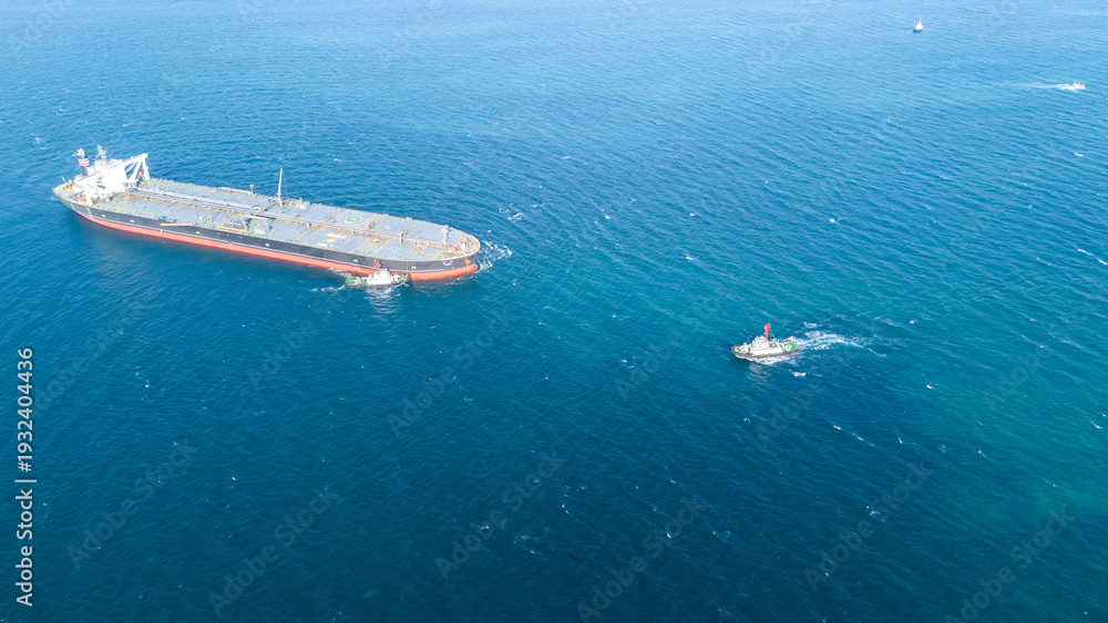 Fototapeta premium Massive crude oil tanker ship assisted by tugboats while sailing across the deep blue ocean. Global maritime logistics, fossil fuel energy, and heavy industrial transport business concept.