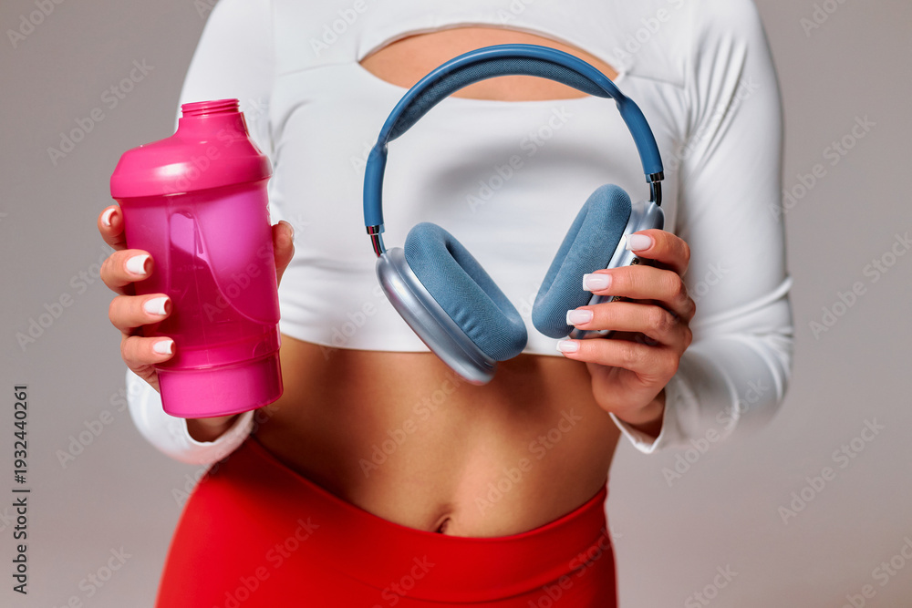 Fototapeta premium Woman holds a pink shaker bottle and blue headphones while standing against a grey background
