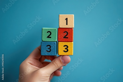 Hand holding five stacked colorful wooden number blocks (1, 2, 3) against a teal background, playful learning and curiosity