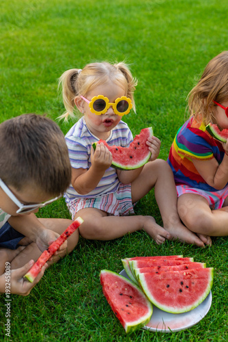 Happy children in sunglasses eating watermelon slices on green grass