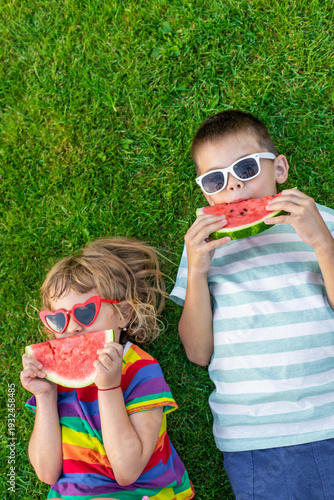 Happy children in sunglasses eating watermelon slices on green grass