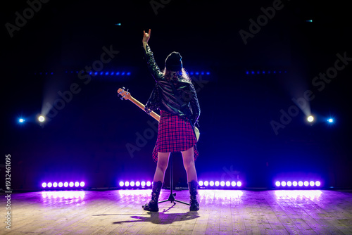 rocker girl in a black leather jacket stands with her back to the camera on stage, raising the sign of the horns hand gesture under stage spotlights.