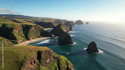 Aerial view of rugged coastline with green hills and rocky cliffs, waves crashing against the shore under clear blue sky during golden hour light