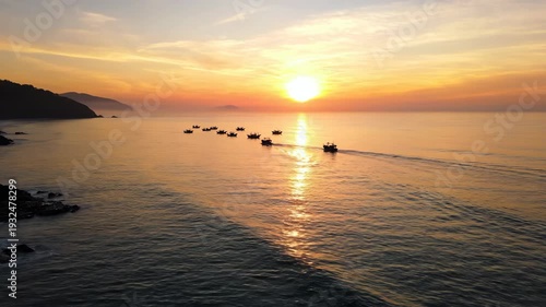 Fishing boats navigate across calm waters at sunset, creating ripples in the ocean as the sun sets behind distant hills, casting warm hues across the sky