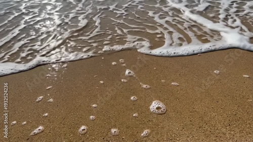 Ocean waves gently wash over sandy beach, creating foam patterns and reflections of sunlight on the water surface, capturing the serene coastal environment