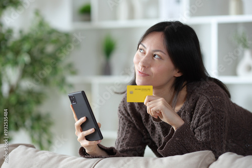 Pensive asian woman holding card and phone at home