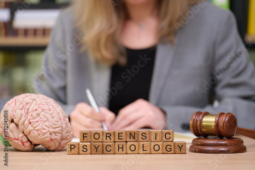 In a modern office, a professional woman engages in research on forensic psychology. Nearby, a brain model, gavel, and letter blocks spell out the focus of her work, showing deep thoughtfulness.
