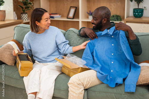 A smiling couple sits together on a green sofa, opening delivery boxes as the man holds up a bright blue shirt, sharing a cheerful moment while shopping online.