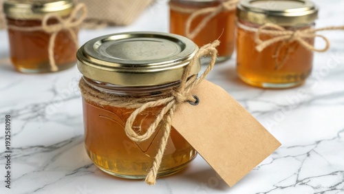 Small glass jars filled with golden honey, tied with twine, featuring a blank craft paper tag for branding or messages, presented on a marble surface.