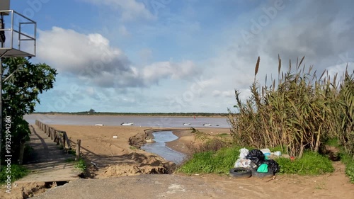 Polluted riverside landscape with a small stream flowing into the river, showing plastic bags and old tires near tall reeds