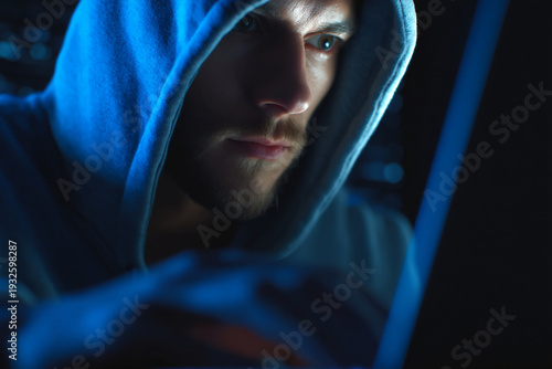 Serious man wearing a blue hoodie is intensely focused on a laptop screen in the dark.