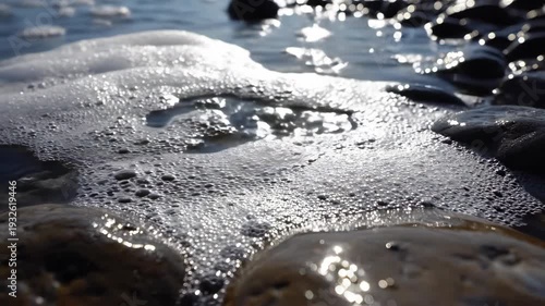Close-up view of water bubbles forming and breaking on smooth stones along a shoreline, sunlight reflecting off the water surface creating a shimmering effect